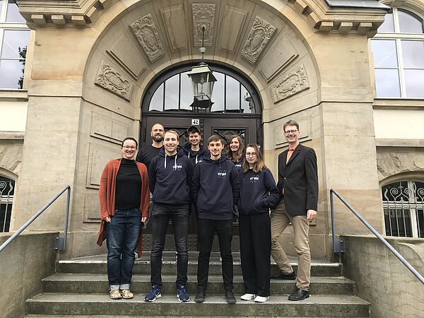 Studentisches Projektteam, sowie Projektmitarbeiter:innen und Professor stehen gemeinsam auf der Treppe vor dem Gutenbergbau.