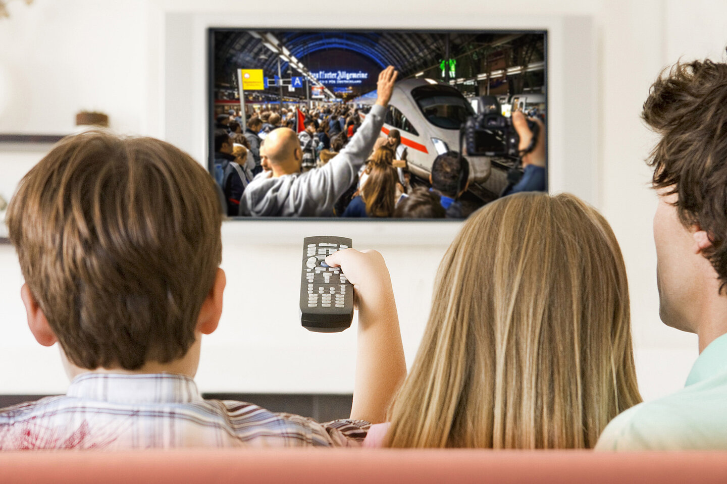 Zwei Kinder vor einem Fernseher, der zeigt, wie im ICE ankommende Geflüchtete von einer Menschenmasse begrüßt werden.