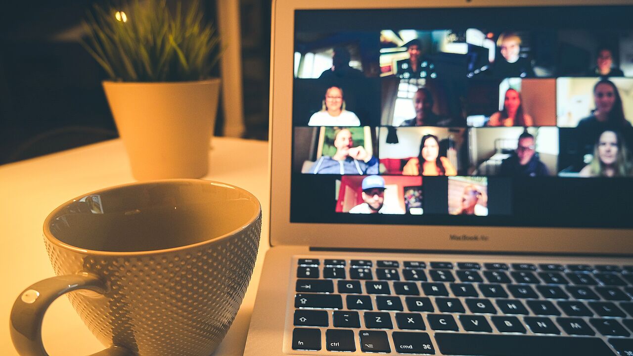 Ein Laptop mit einer laufenden Online-Konferenz auf dem Bildschirm steht neben einer Tasse.