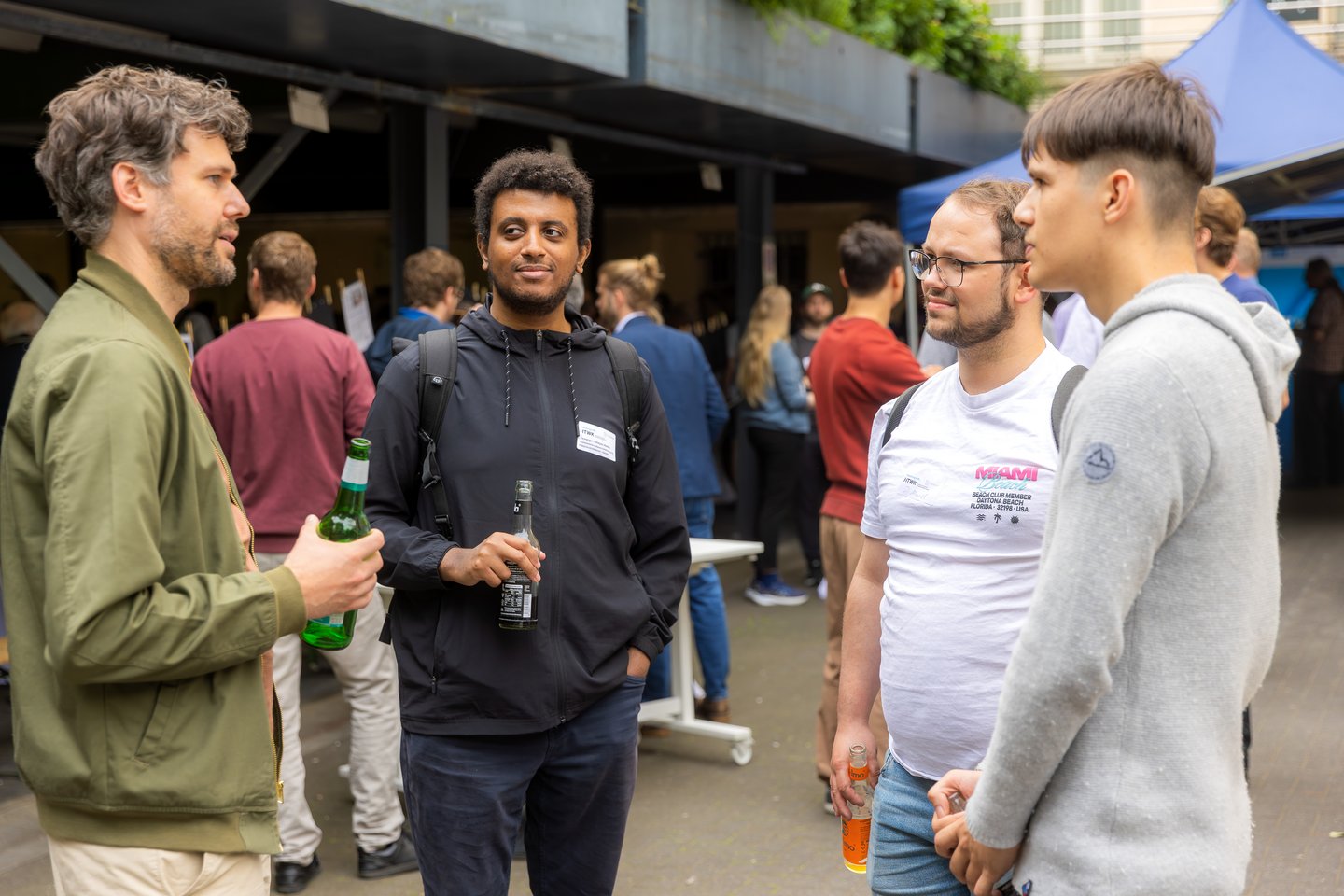 Forschung trifft ... 2026 Vier Männer stehen nebeneinander, unterhalten sich und halten jeweils ein Getränk in der Hand. Im Hintergrund sind weitere Gruppen von Menschen zu sehen