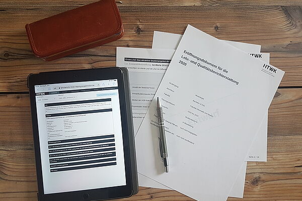 Auf einem Tisch liegt ein Tablet mit geöffnetem digitalen Lehrbericht. Daneben liegen Ausdrucke einiger Berichtsseiten und ein Stift.