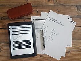 Auf einem Tisch liegt ein Tablet mit geöffnetem digitalen Lehrbericht. Daneben liegen Ausdrucke einiger Berichtsseiten und ein Stift.